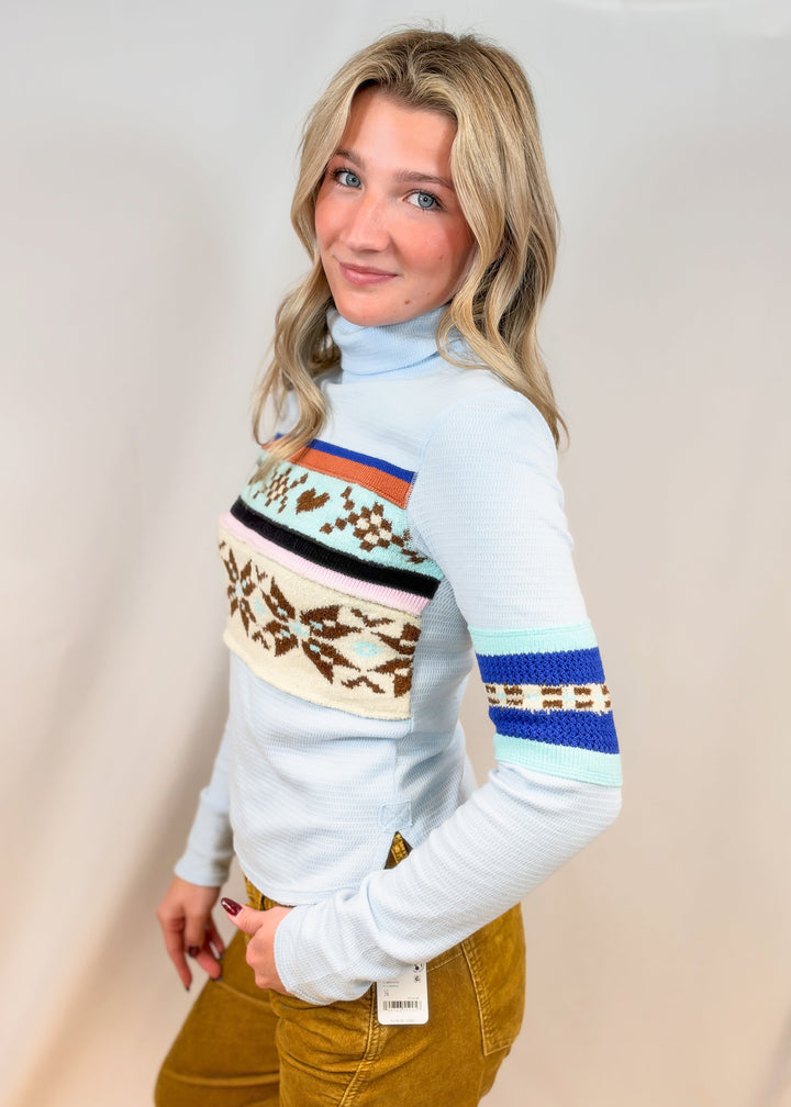 Woman shown from side wearing light blue turtle neck top with argyle and snowflake designs with camel cord pants on plain background