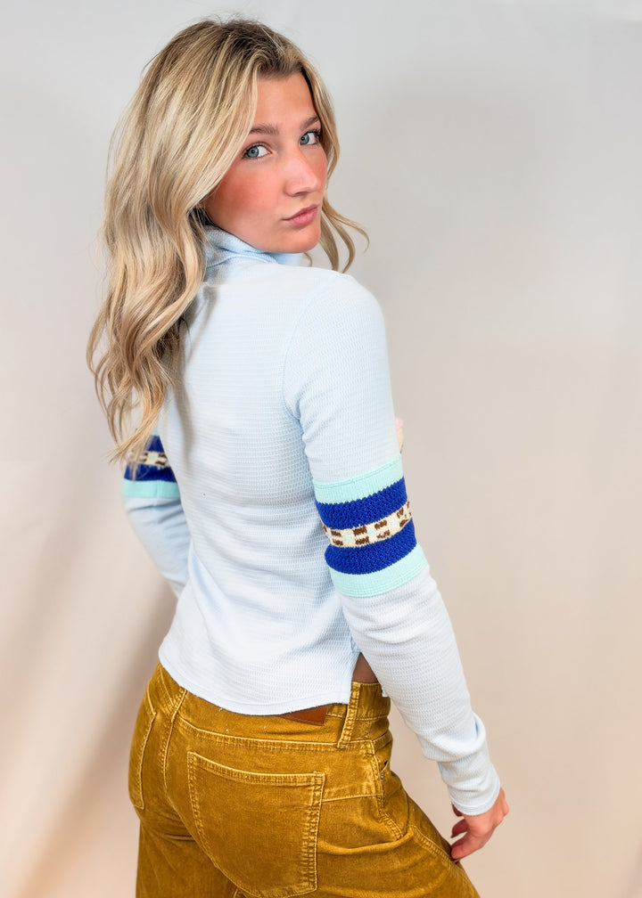 Woman shown from side view wearing light blue turtleneck top with argyle and snowflake designs with camel cord pants on plain background