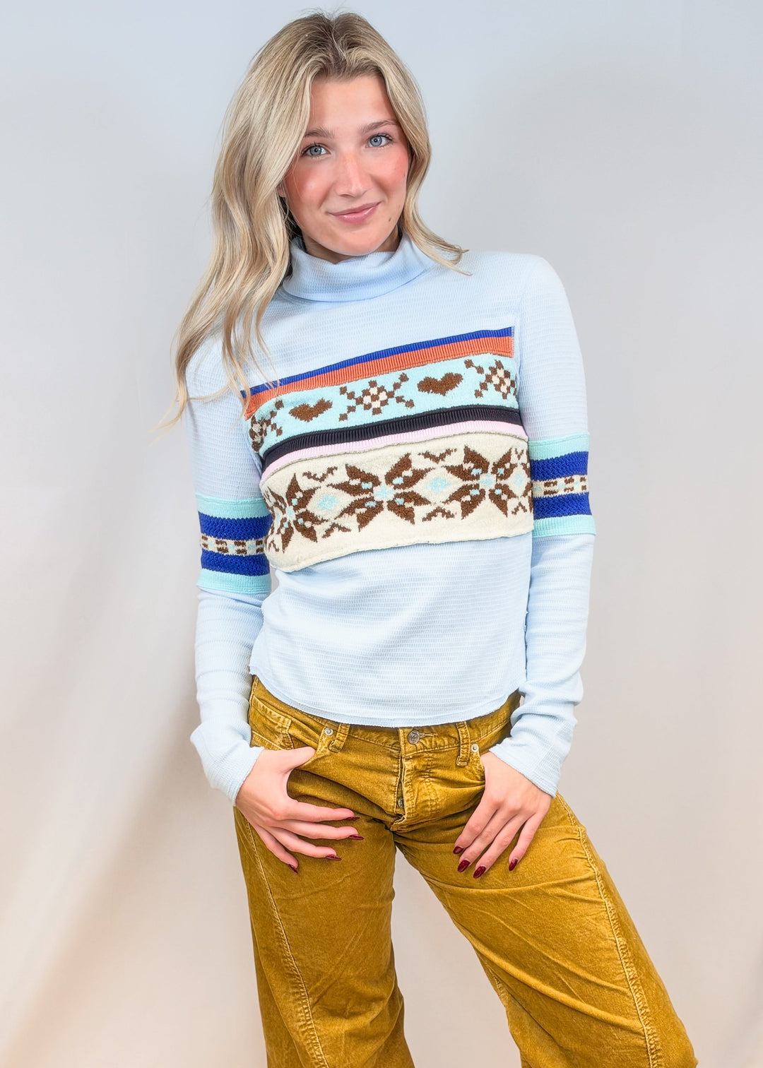 Woman wearing light blue turtleneck top with argyle and snowflake designs with camel cord pants on plain background