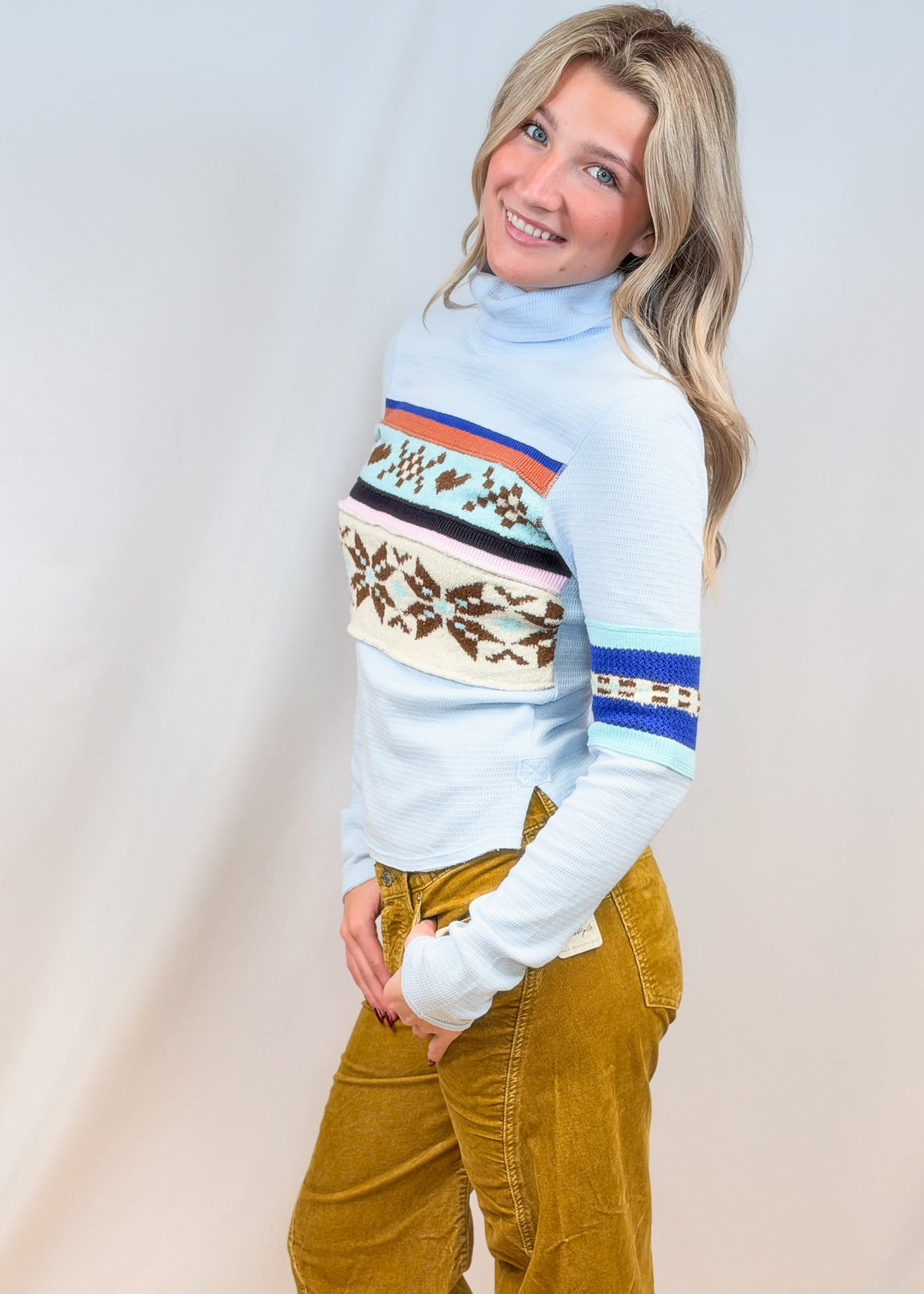 Woman shown from side view wearing light blue turtleneck top with argyle and snowflake designs with camel cord pants on plain background