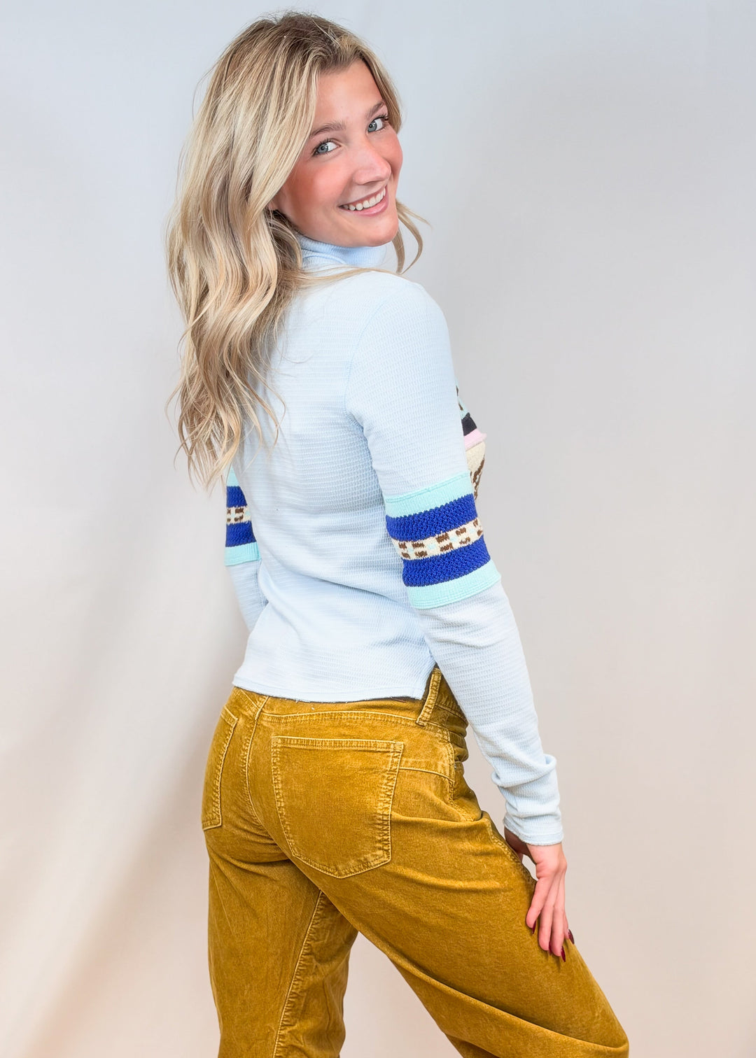 Woman shown from side view wearing light blue turtleneck and camel cord pants on plain background