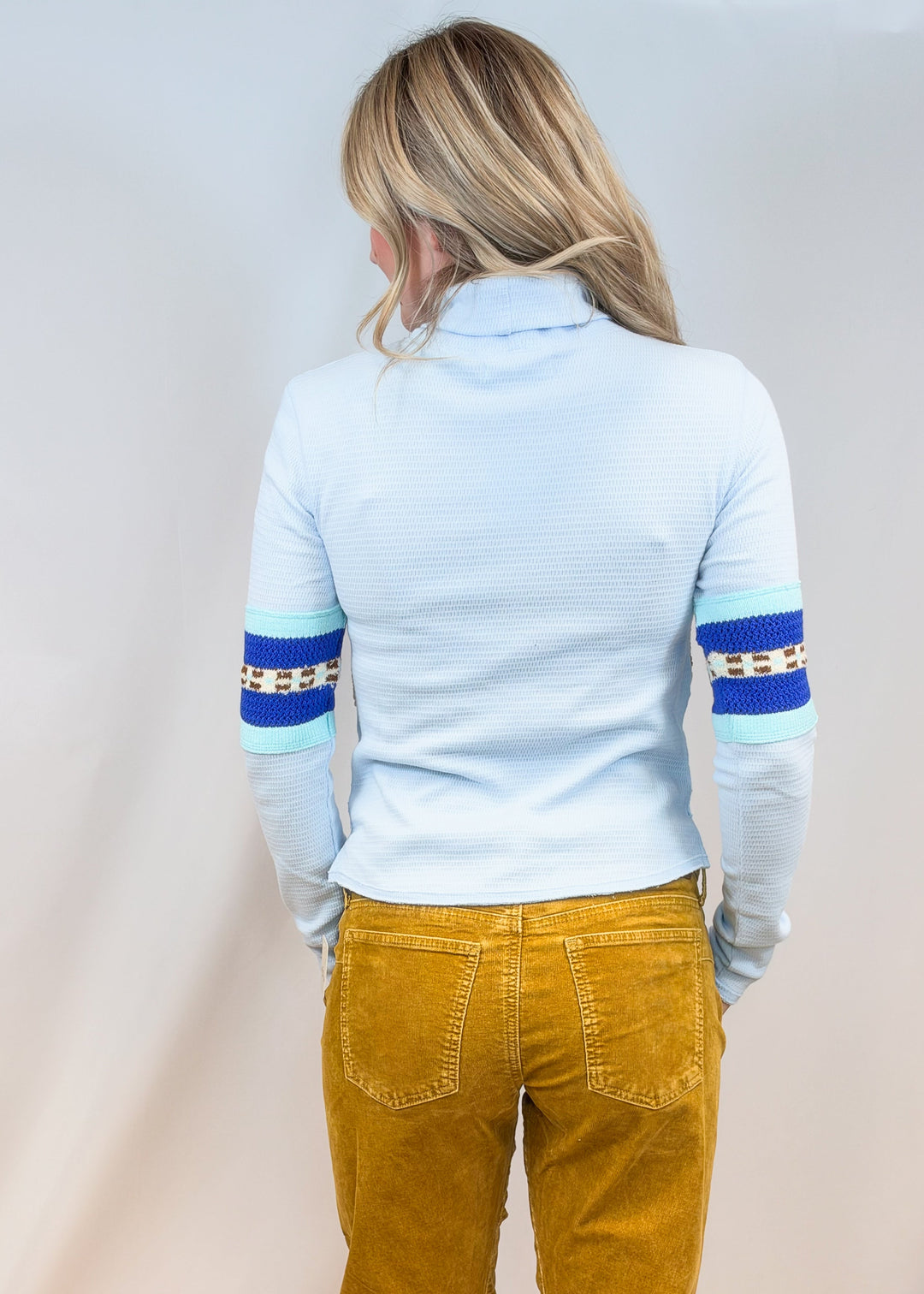 Woman shown from back view wearing light blue turtleneck and camel cord pants on plain background
