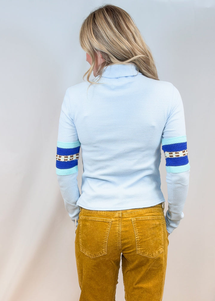 Woman shown from back view wearing light blue turtleneck and camel cord pants on plain background