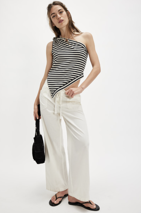 Woman wearing a black and white striped sleeveless top and white pants, holding a black handbag on a white background.