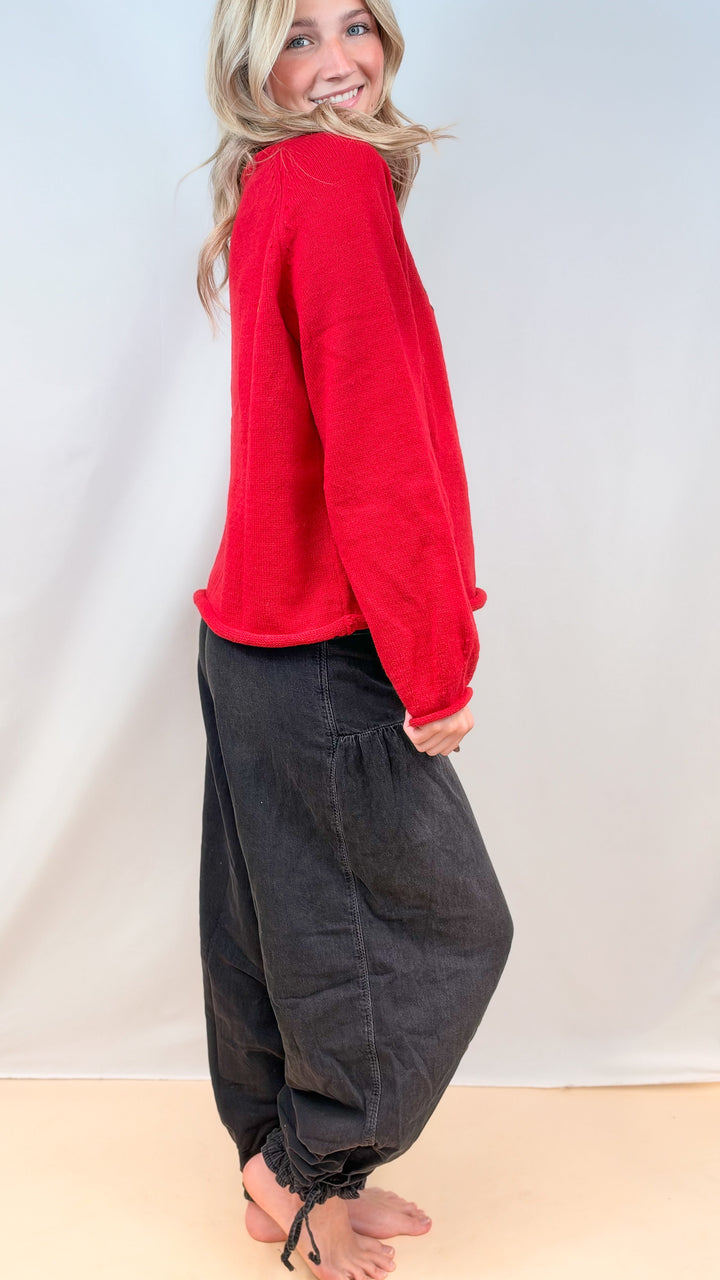 Woman shown from side view wearing red sweater and black balloon pants on plain background
