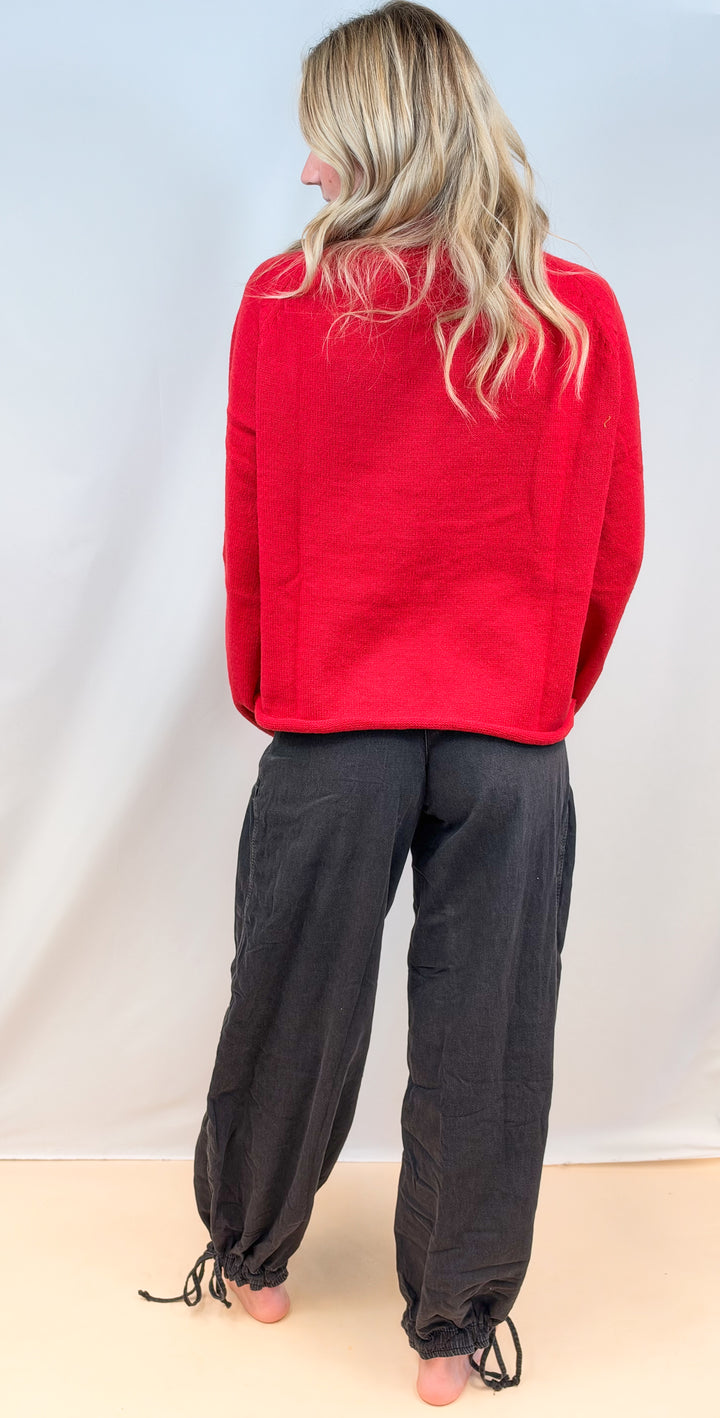 Woman shown from back view wearing red sweater and black balloon pants on plain background