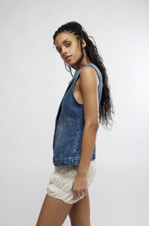 Woman wearing a denim vest over a white top and shorts on a plain background