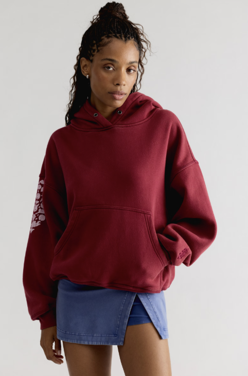 Woman wearing red hooded sweatshirt with logo on sleeve with blue skirt on plain background