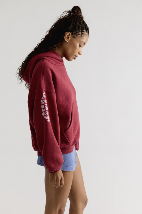 Woman shown from side view wearing red hooded sweatshirt with logo on sleeve with blue skirt on plain background
