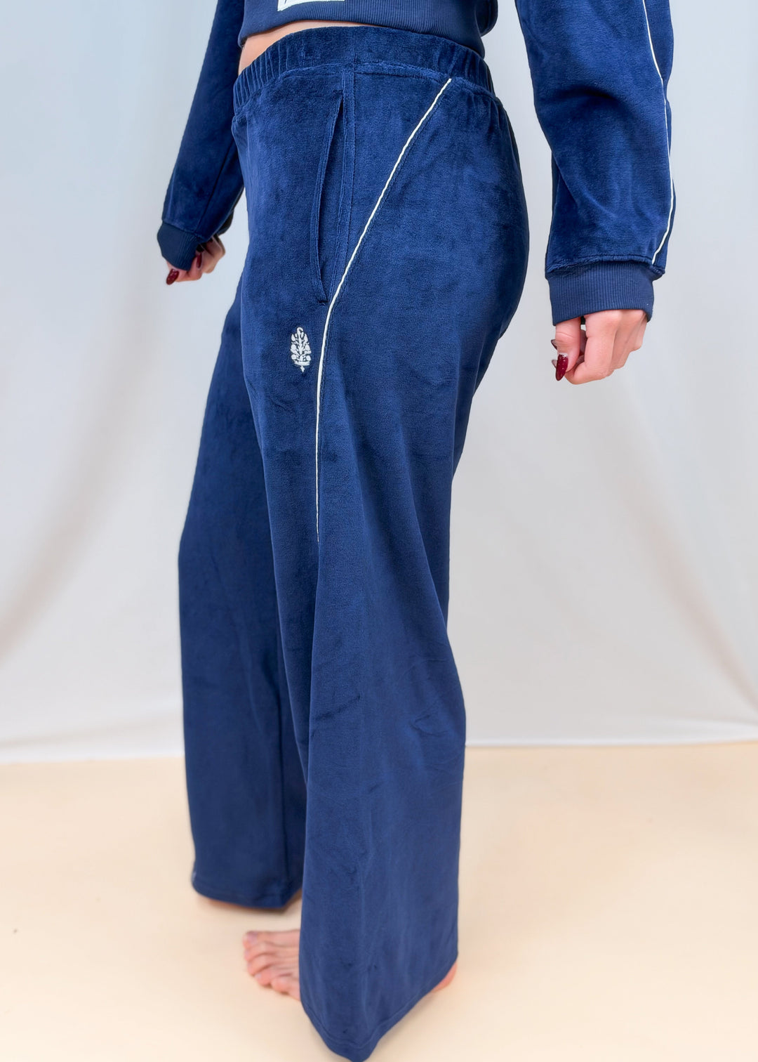 Woman shown from side view wearing blue velour pants with white piping and brand logo on plain background