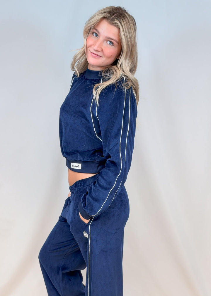 Woman shown from side view wearing blue matching velour set with white piping on plain background