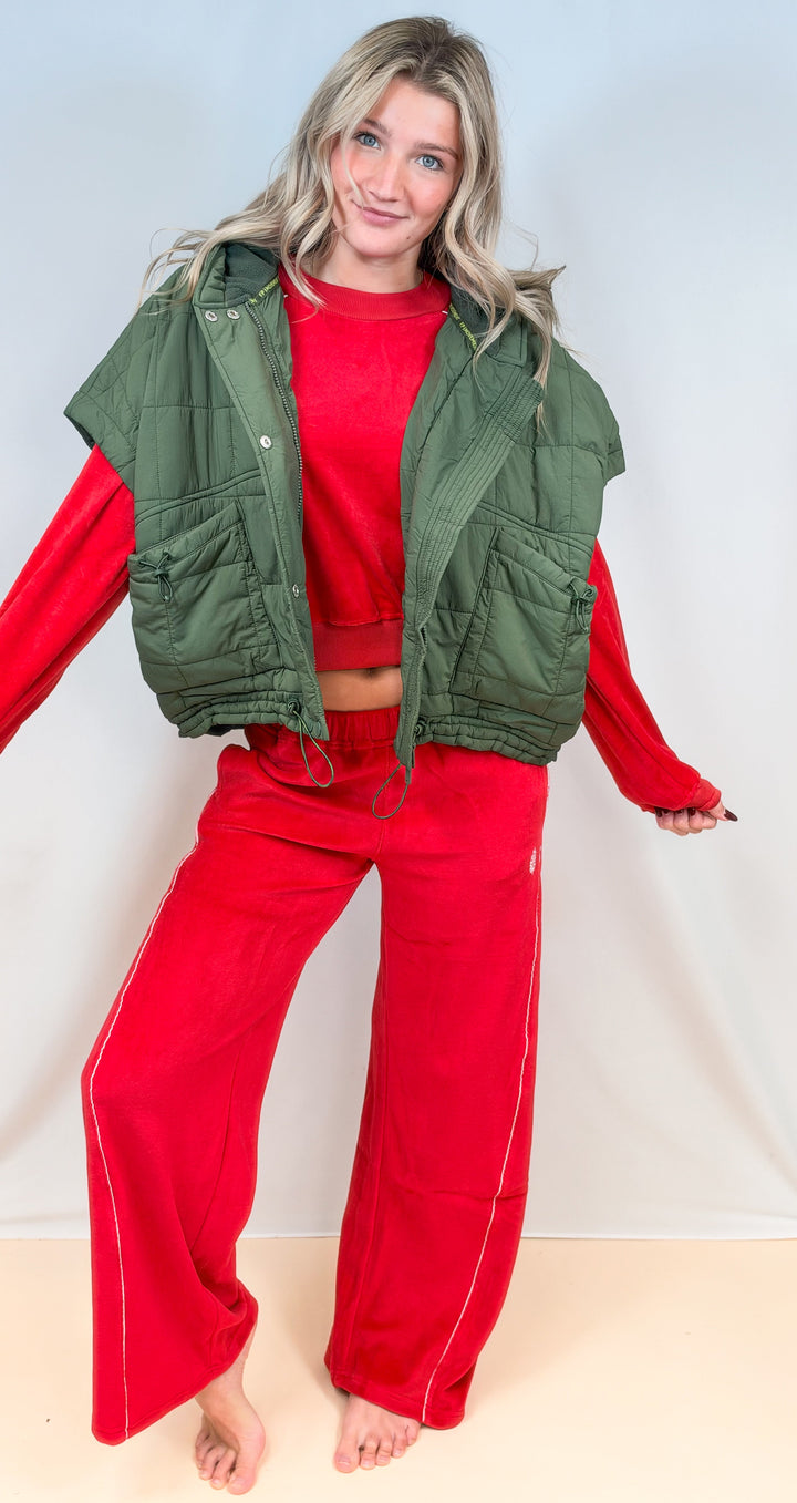 Woman wearing matching red velour set with puffy quilted olive vest on plain background