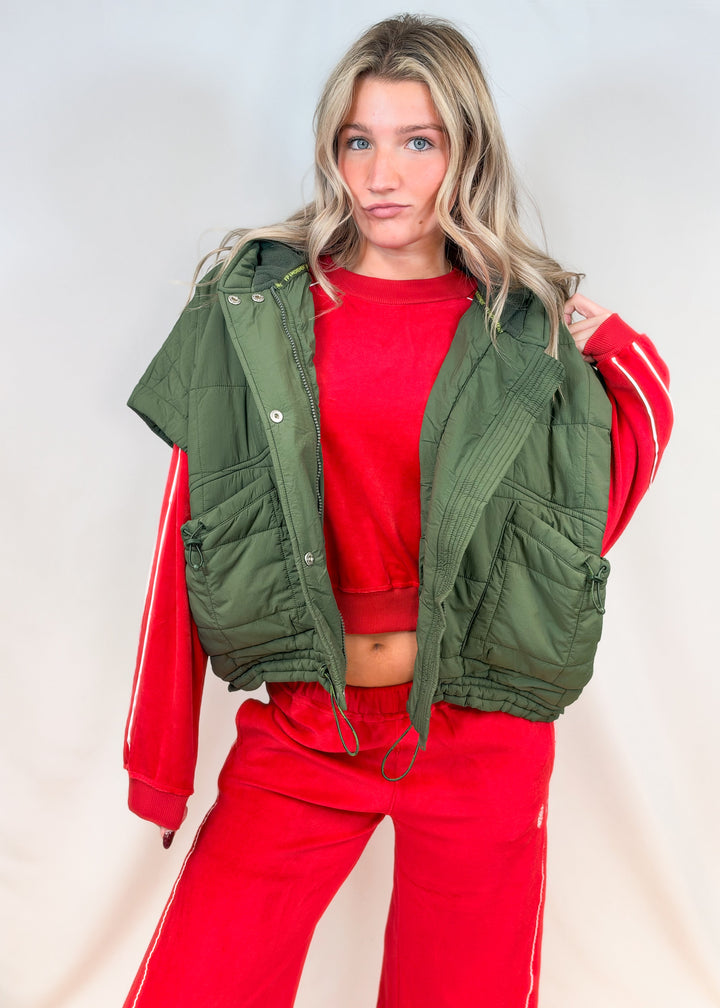 Woman wearing red matching velour set with olive green puffy quilted vest on plain background