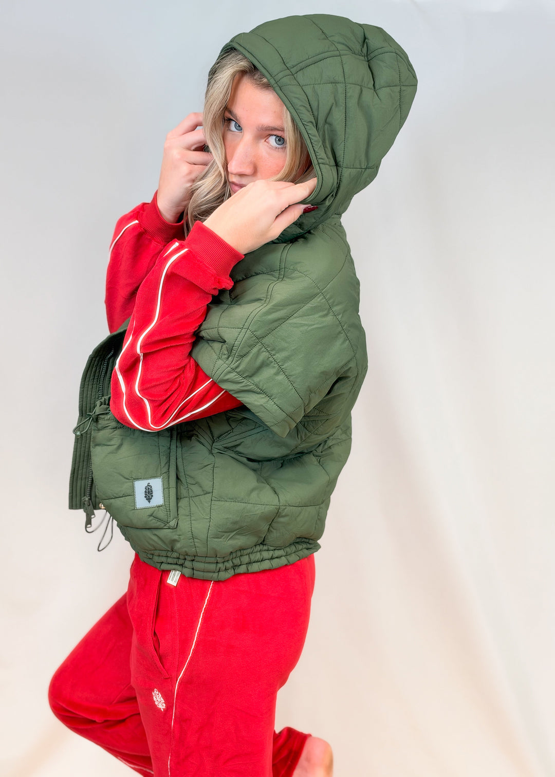Woman shown from side view wearing matching red velour set with olive green quilted puffy vest on plain background