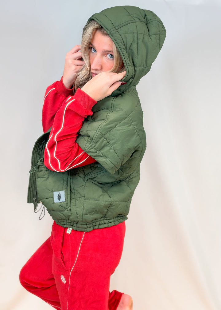 Woman shown from side view wearing matching red velour set with olive green quilted puffy vest on plain background