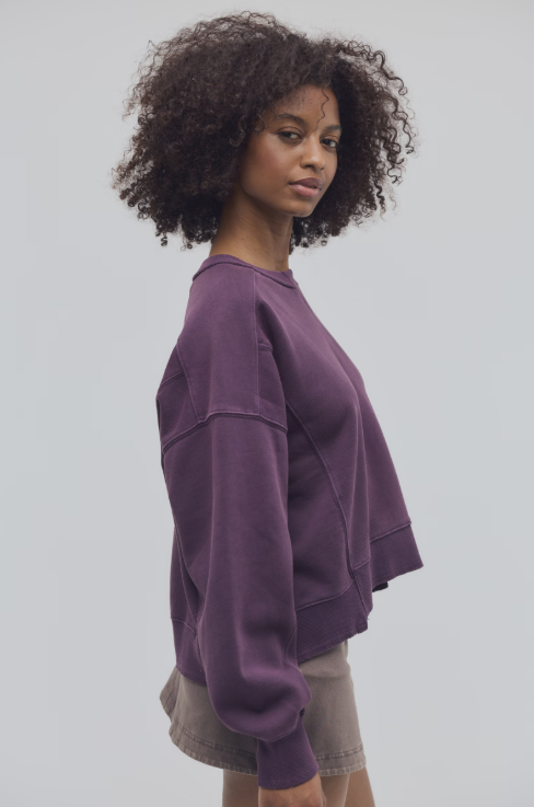 Woman shown from side view wearing purple sweatshirt and brown skirt on plain background