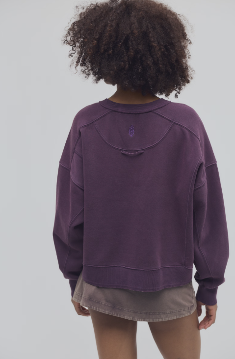 Woman shown from back view wearing purple sweatshirt and brown skirt on plain background