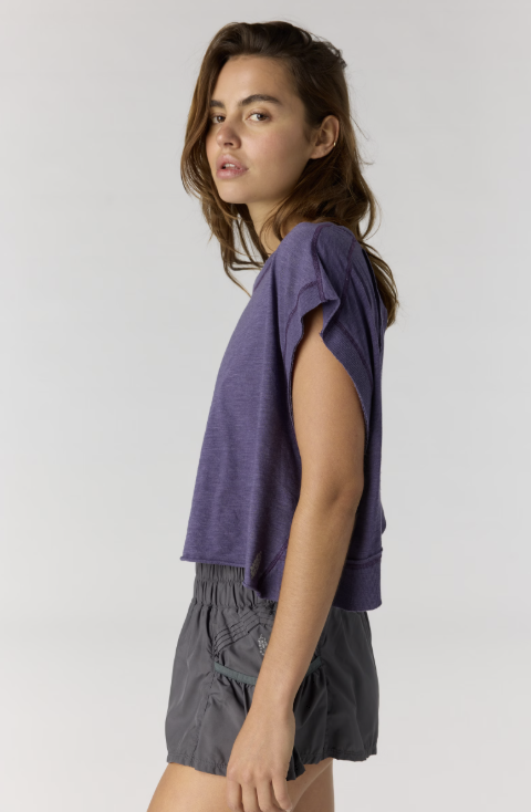 Woman shown from side view wearing purple short sleeve shirt and black shorts on plain background