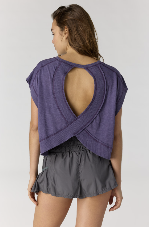 Woman shown from back view wearing purple short sleeve top with back cut out and black shorts on plain background
