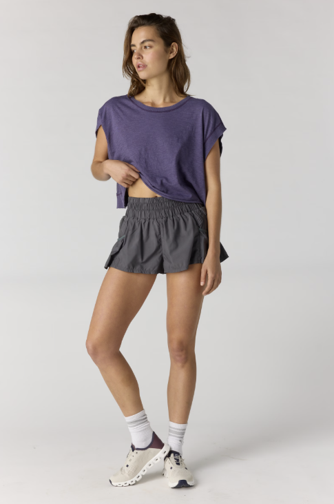 Woman wearing purple short sleeve top with black shorts and sneakers on plain background
