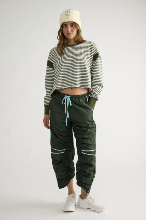 A model stands against a light gray studio background wearing a green and white striped cropped long-sleeve top, dark green cargo-style pants with a light blue drawstring and white piping, a cream beanie, and white sneakers.