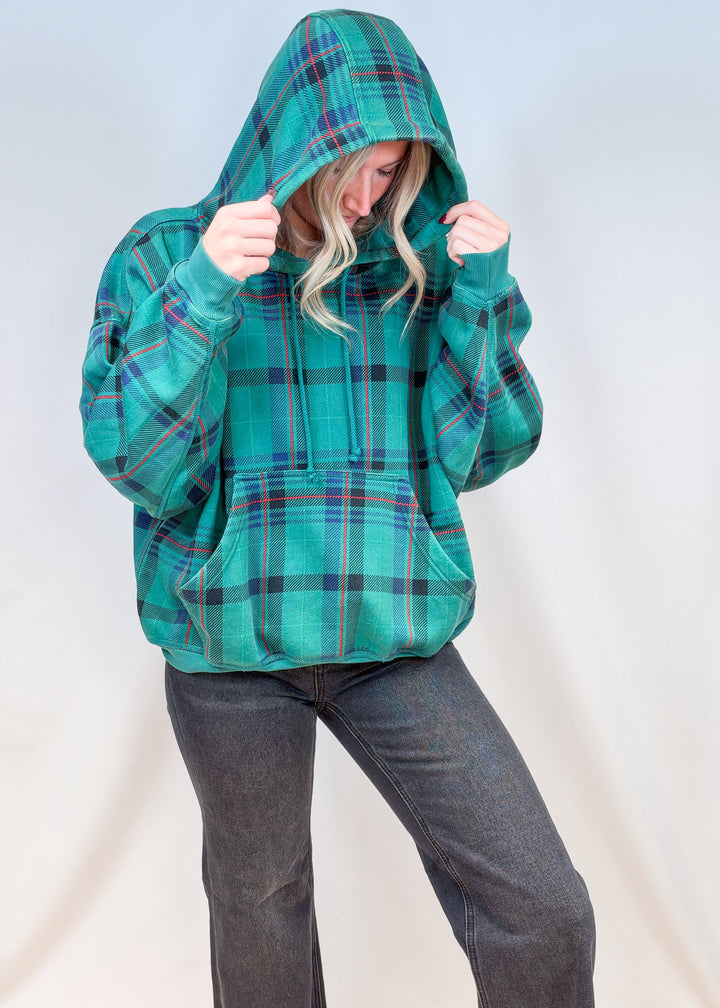 Woman wearing green and blue plaid hoodie with black pants on plain background