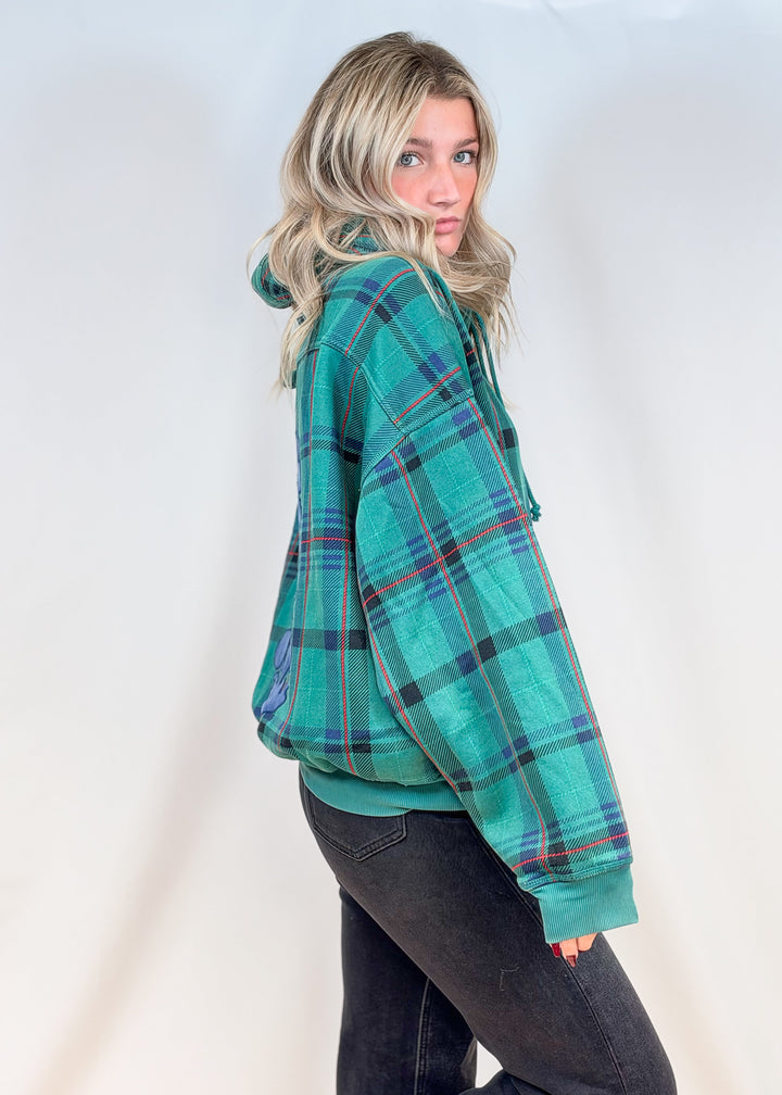 Woman shown from side view wearing green and blue plaid hoodie with black pants on plain background