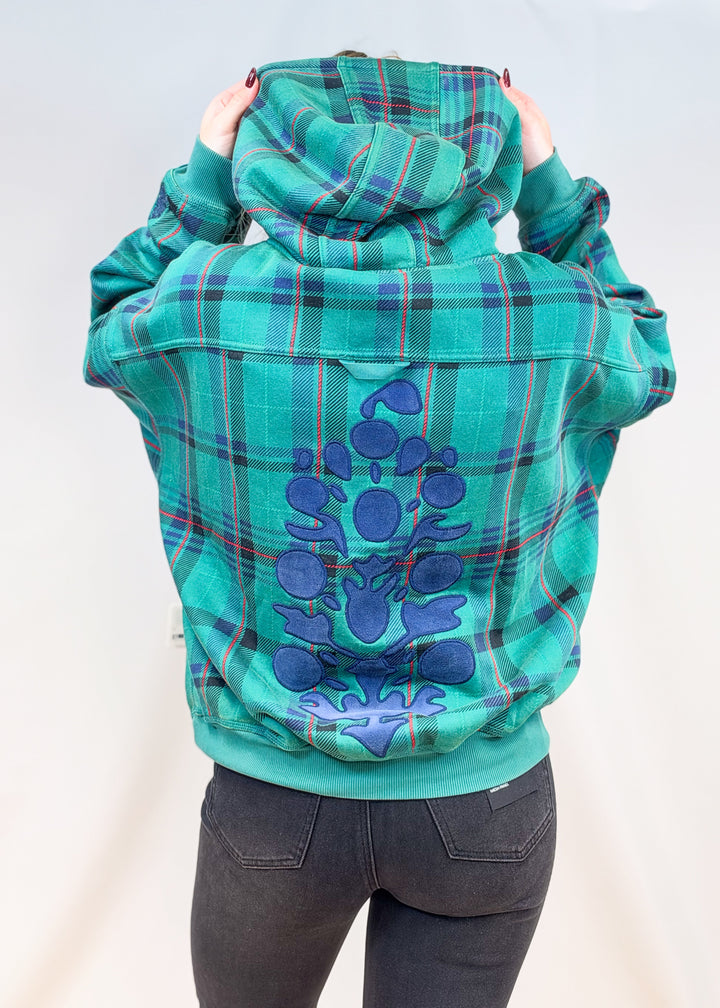 Woman shown from back view wearing green and blue plaid hoodie with large blue decal in velour on back with black jeans on plain background