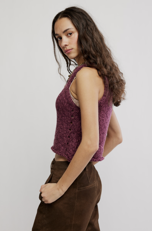 A woman modeling a sleeveless, textured knit vest in a deep plum purple color, paired with brown pants, against a plain light gray studio background.