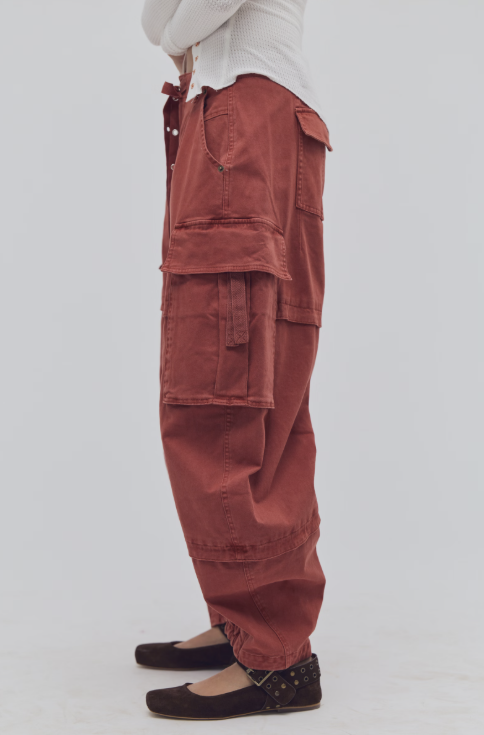 Woman shown from side view waist down wearing white top and red rust color cargo pants with flats on plain background
