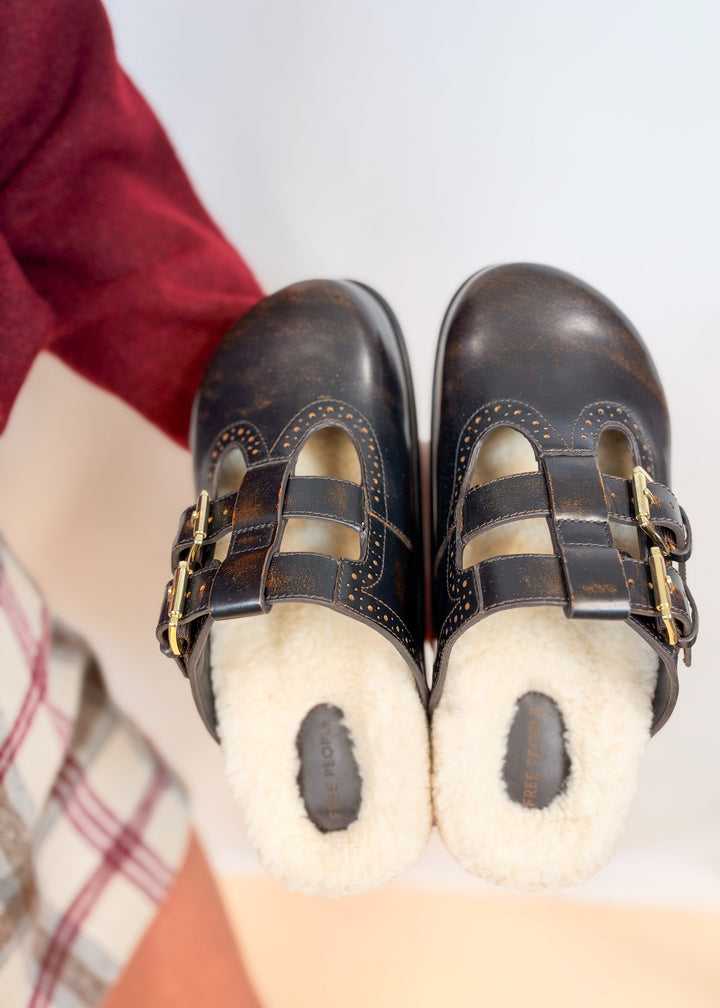Person holding brown leather mule shoes with shearling inner with plain background