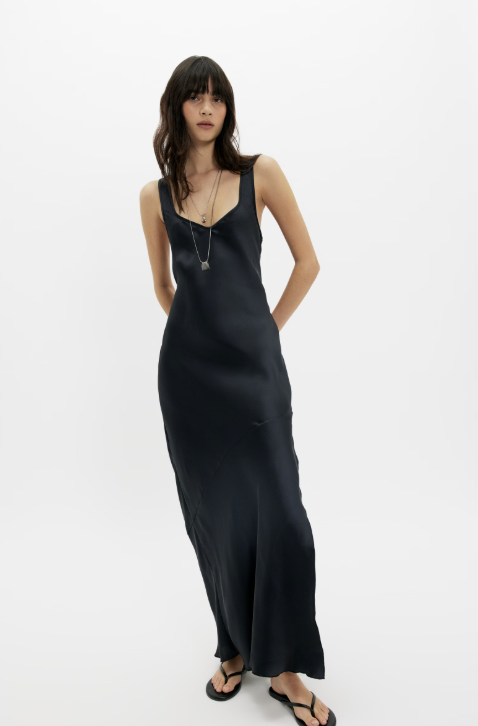 A woman modeling a long black satin slip dress with a deep V-neck and thin straps, paired with black sandals and a silver pendant necklace, set against a plain white studio background.