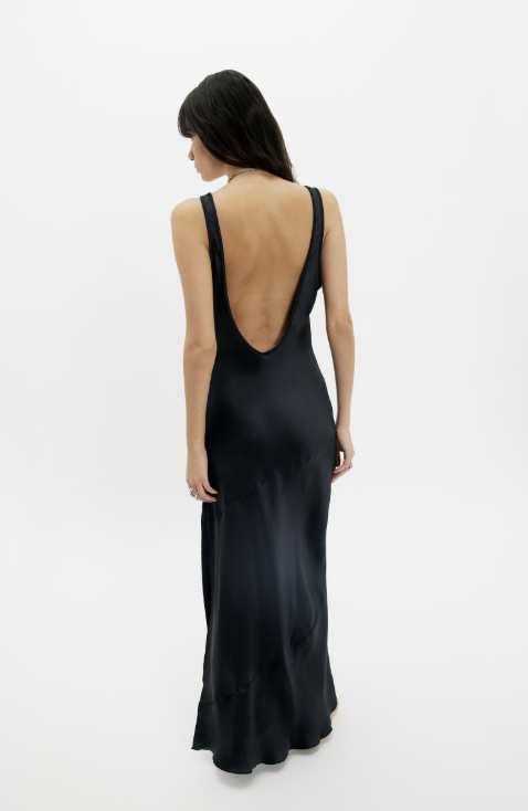 A woman viewed from the back wearing a black satin maxi dress with a deep open back, photographed against a clean white studio background.