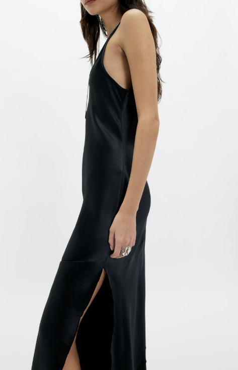 A woman wearing a black satin slip dress with a high side slit, shown in a side profile against a plain white studio background.