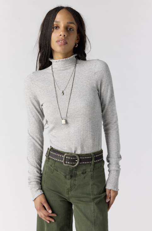Woman wearing grey body fitting mock neck top with green pants on plain background
