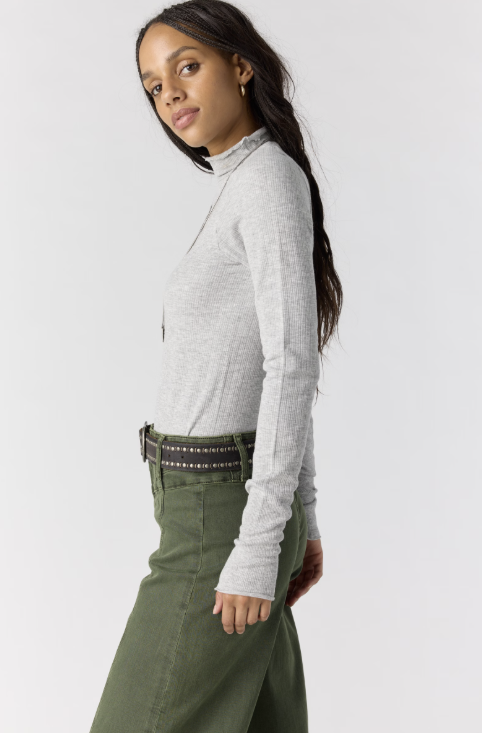Woman shown from side view wearing grey body fitting mock neck top and green pants on plain background
