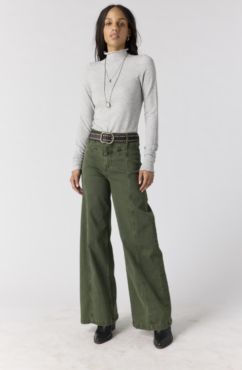 Woman wearing grey body fitting mock neck top with green pants on plain background