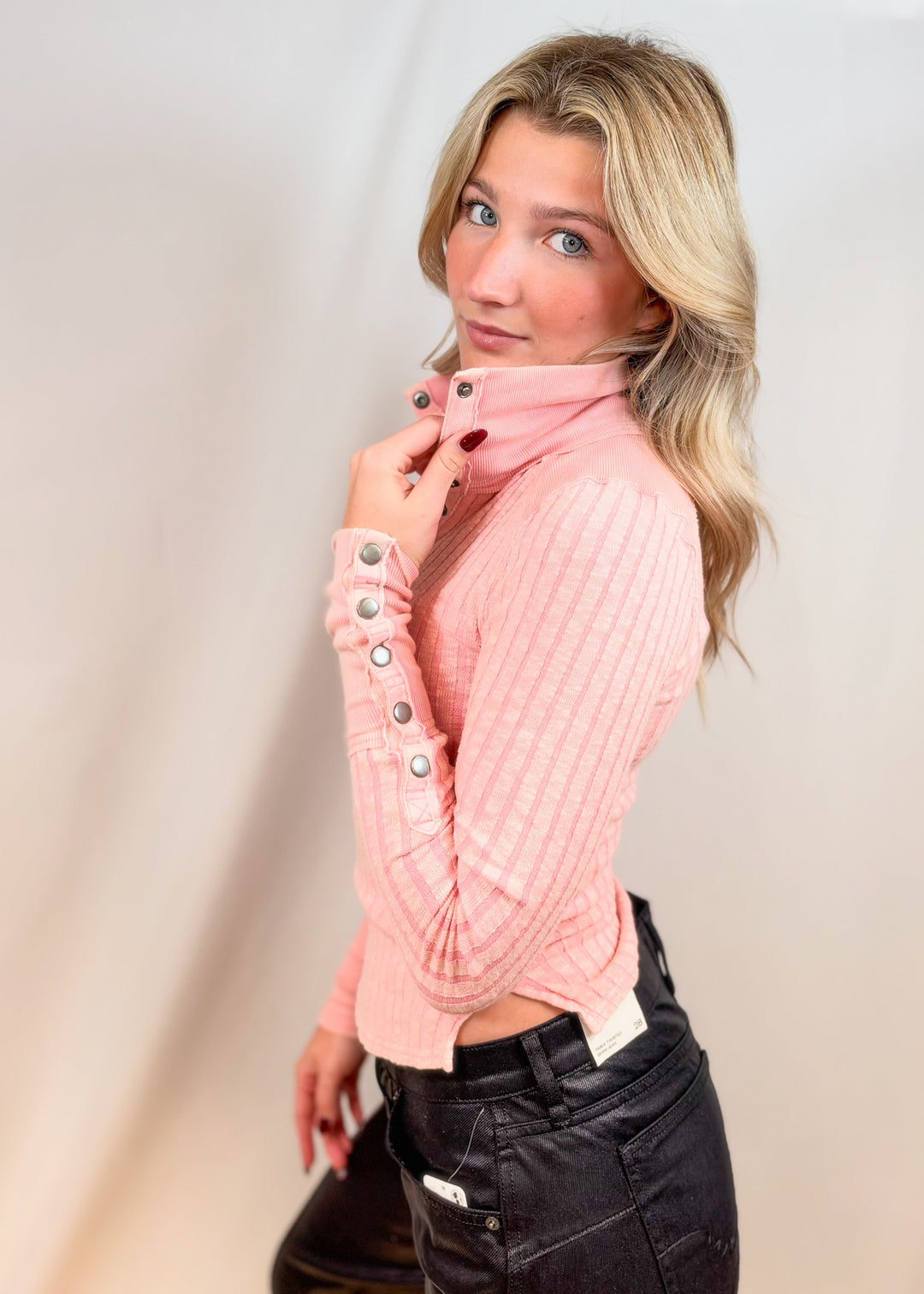 Woman wearing pink henley top with button cuffs and black pants on plain background