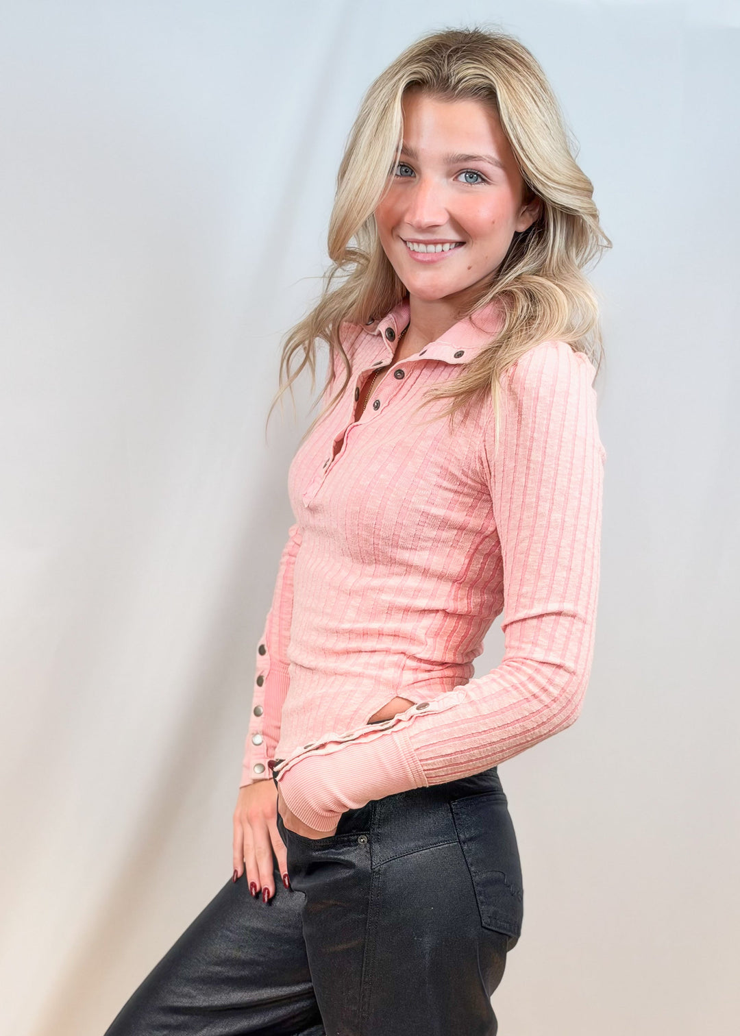 Woman wearing pink henley with botton collar and cuffs with black pants on plain background