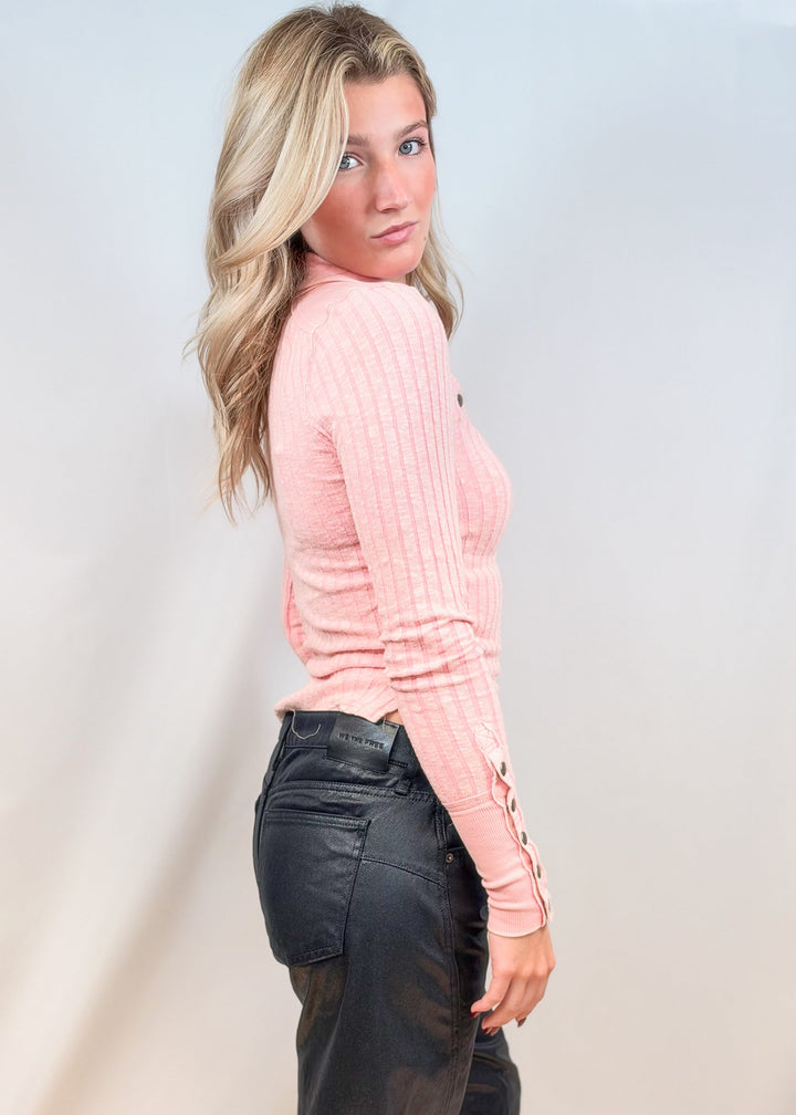 Woman wearing pink henley with black pants on plain background