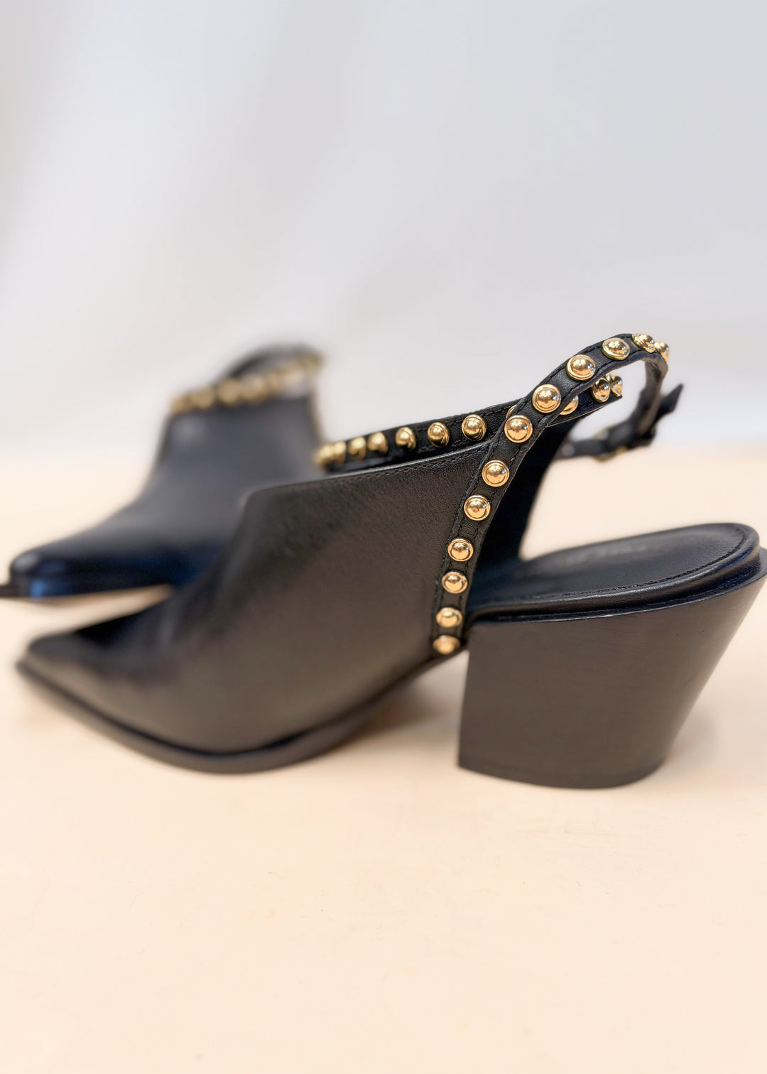Pair of black leather pointed toe heels with gold studded leather wraps on plain background