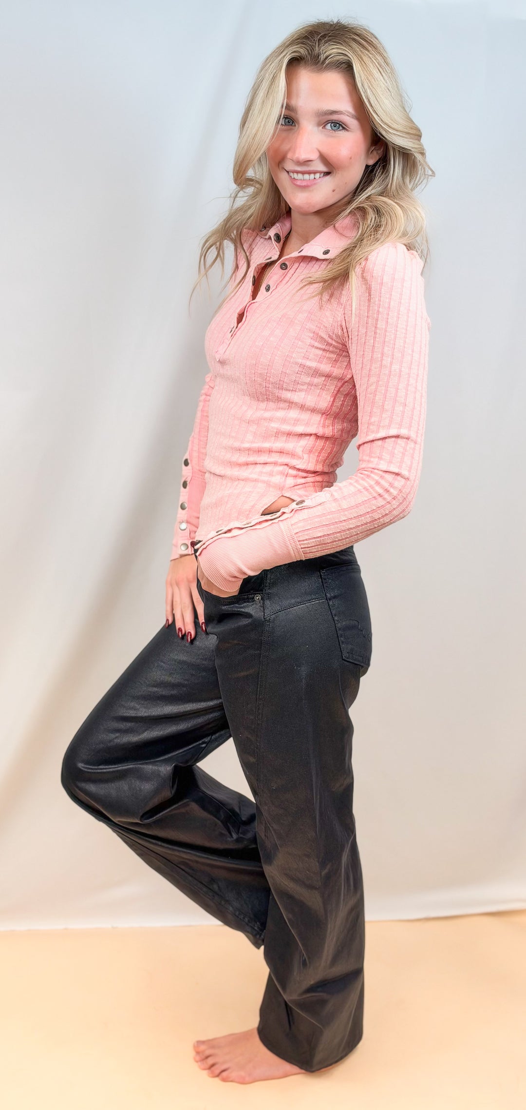 Woman shown from side view wearing pink henley and black pants on plain background