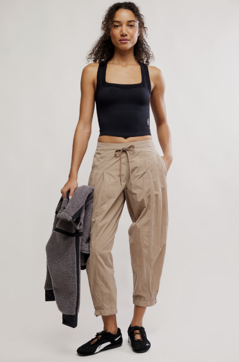 Woman wearing a black tank top and beige pants holding a gray jacket on a white background