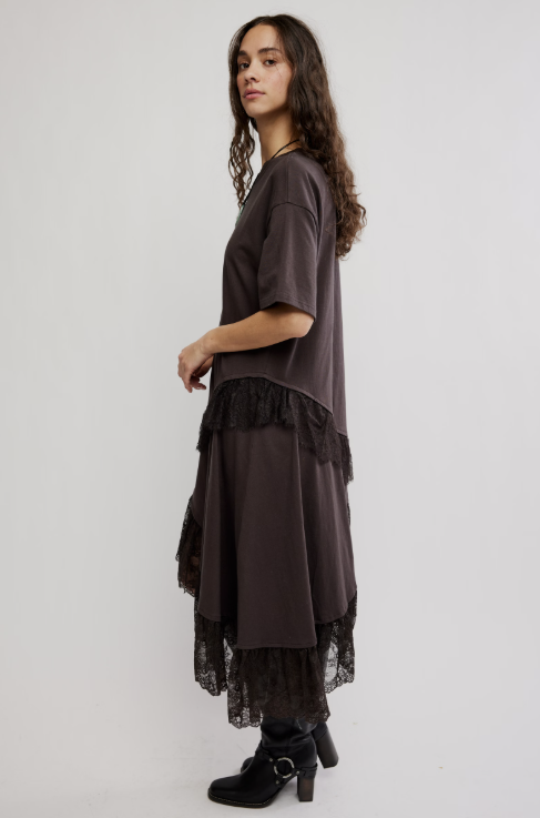 A woman modeling a dark brown short-sleeve tunic dress with black lace ruffle trim, paired with black ankle boots, against a plain light gray studio background.