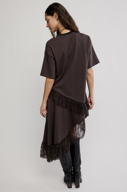 A woman shown from the back wearing an oversized dark brown short-sleeve top with a black lace ruffle trim at the hem, paired with dark pants and black ankle boots, against a plain light gray studio background.