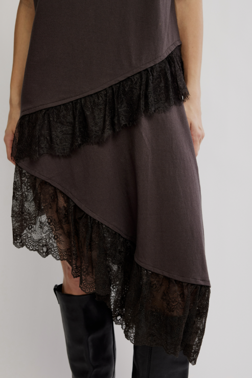 A close-up of a model wearing a dark brown asymmetrical dress with layered black lace ruffles and black knee-high boots against a light gray studio background.