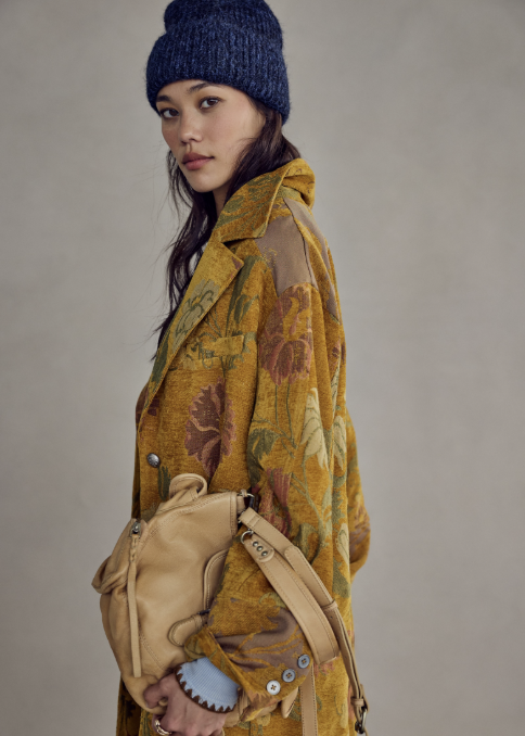A model is shown in profile wearing a mustard yellow floral patterned coat, a navy blue knit beanie, and carrying a tan leather shoulder bag against a neutral gray studio background.