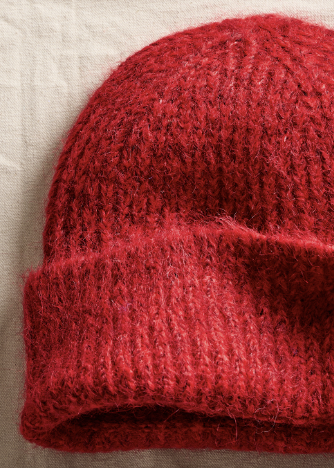 A close-up of a red fuzzy knit beanie hat resting on a light beige fabric background.
