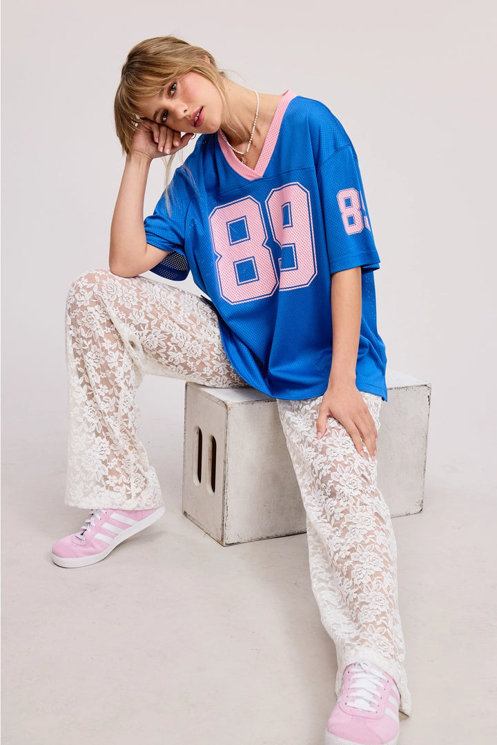 Woman wearing a blue oversized jersey with the number 89, white lace pants, and pink high-top sneakers on a light gray background.