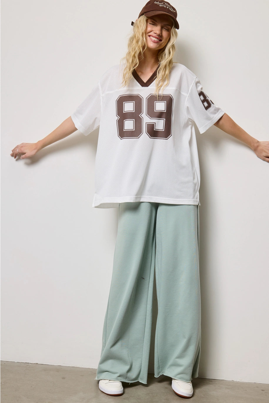 Person wearing a white oversized jersey with brown numbers and green pants against a plain background